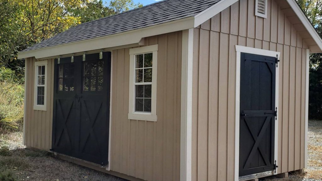 Tan storage shed for sale with two windows, front and side door entrances, and a gray shingle roof